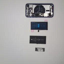 Rozłożony iPhone 12 z widocznymi komponentami: obudowa, dwie baterie i moduł pamięci, ułożone na jasnym tle.