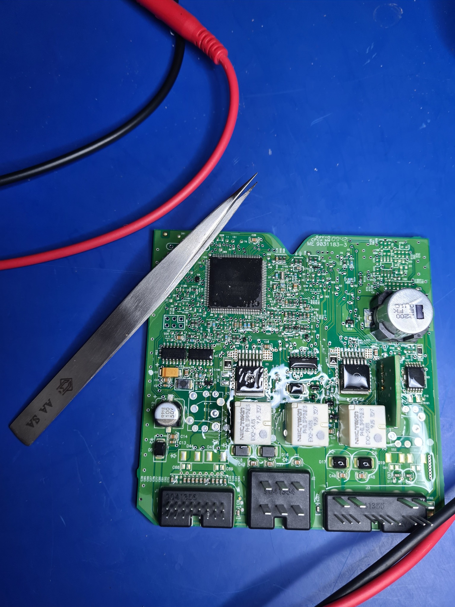 Zielona płytka drukowana z elementami elektronicznymi poddawana inspekcji przy użyciu pęsety, widoczne przewody multimetru na niebieskim tle.