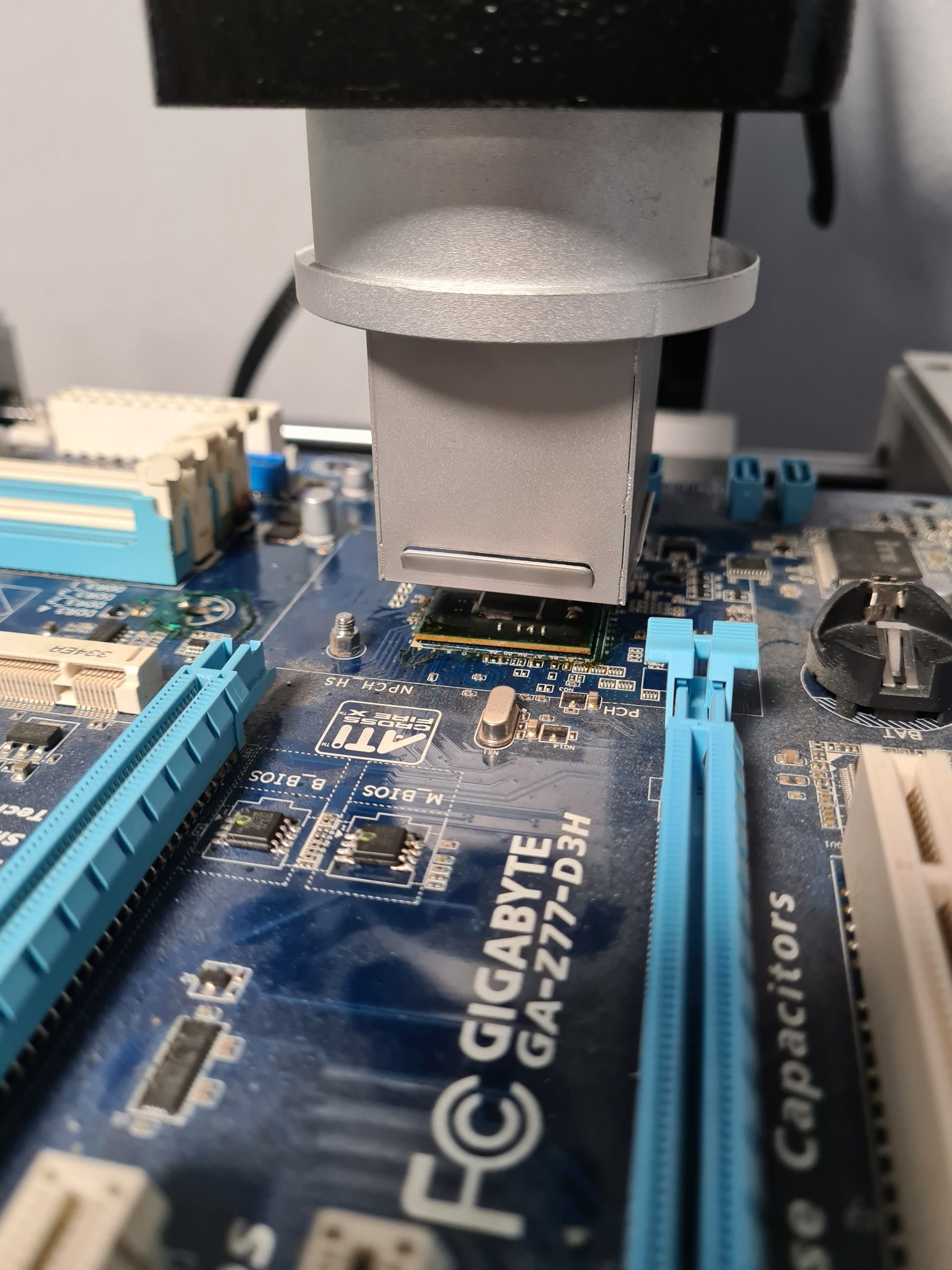Szczegółowe ujęcie płyty głównej Gigabyte GA-Z77-D3H poddawanej naprawie, z użyciem specjalistycznego narzędzia lutowniczego precyzyjnie umieszczonego nad chipem, widoczne niebieskie sloty RAM...