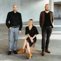 Zespół Fine Marketing: kobieta siedząca na drewnianym stołku pomiędzy dwoma mężczyznami w nieformalnych strojach na tle surowego, betonowego wnętrza.