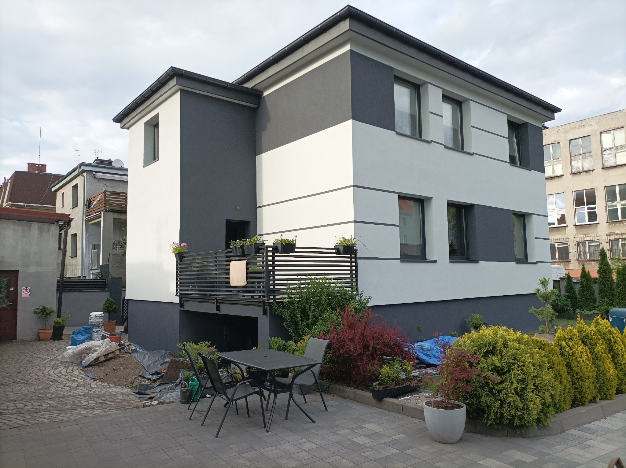 Dwukondygnacyjny dom z elewacją w pasy szaro-białe, balkon z kwiatami, meble ogrodowe na brukowanym podwórku, widoczne prace ziemne.