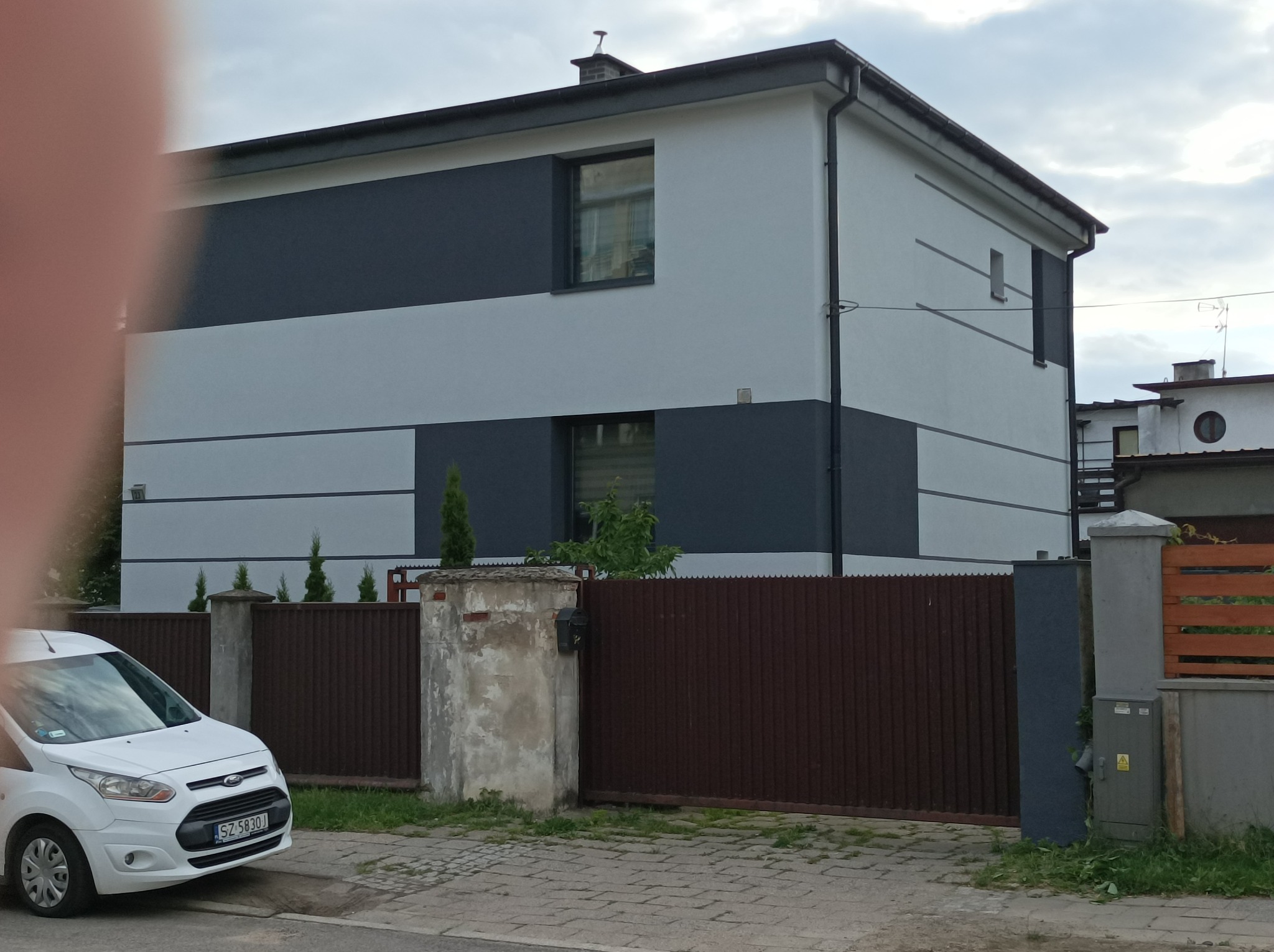 Dwukolorowa elewacja domu jednorodzinnego z poziomymi pasami szarości i bieli, widoczny fragment białego samochodu i brązowe ogrodzenie