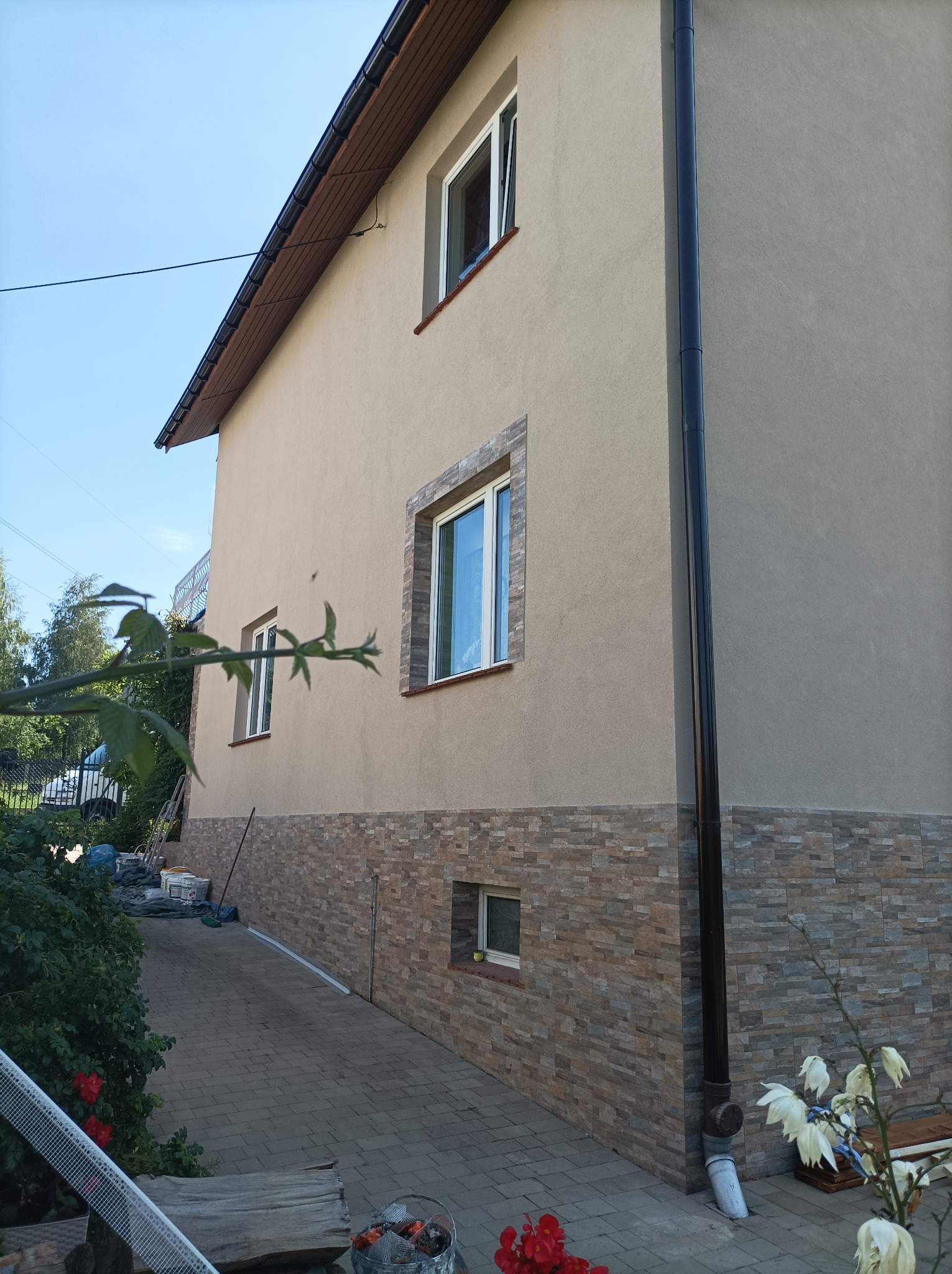 Dwupiętrowy dom z beżową elewacją i kamienną okładziną na dole, widok z boku z fragmentem chodnika i roślinności w kadrze.