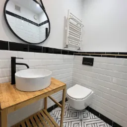 Nowoczesna łazienka w Bolesławcu z białą ceramiką, umywalką nablatową na drewnianym stelażu, czarną armaturą i geometryczną posadzką w czarno-białe wzory.