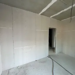 Wnętrze w trakcie prac wykończeniowych: ściany obłożone płytami gipsowo-kartonowymi, widoczne szpachlowane łączenia na suficie, surowy beton na podłodze, wystające kable elektryczne.