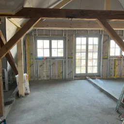 Poddasze w trakcie adaptacji: widoczne drewniane krokwie, metalowy stelaż pod okładzinę, izolacja z folią paroizolacyjną i częściowo zamontowane płyty gipsowo-kartonowe na suficie.