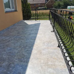 Wyłożony płytkami taras z czarną, metalową balustradą, widok na ogród i zabudowania w tle.