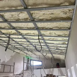 Ocieplenie poddasza pianą PUR widoczne przez metalowy stelaż, na ścianie widoczna poziomica laserowa, w tle okno i rusztowanie.