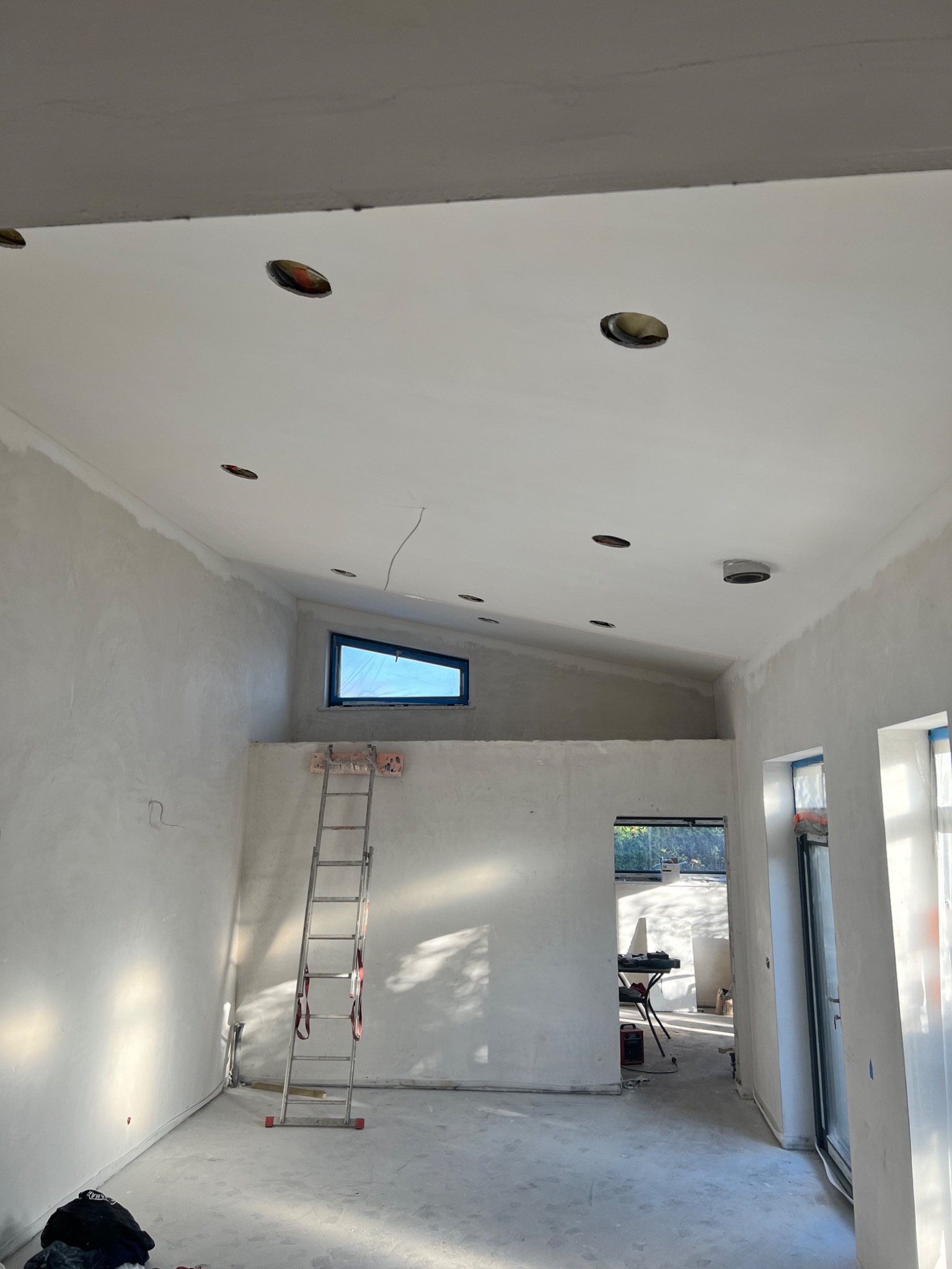 Wnętrze w trakcie prac wykończeniowych: surowe ściany po nałożeniu gładzi, przygotowane otwory pod oświetlenie w suficie, drabina oparta o ścianę pod skośnym oknem, widok na kolejne pomieszczenie.