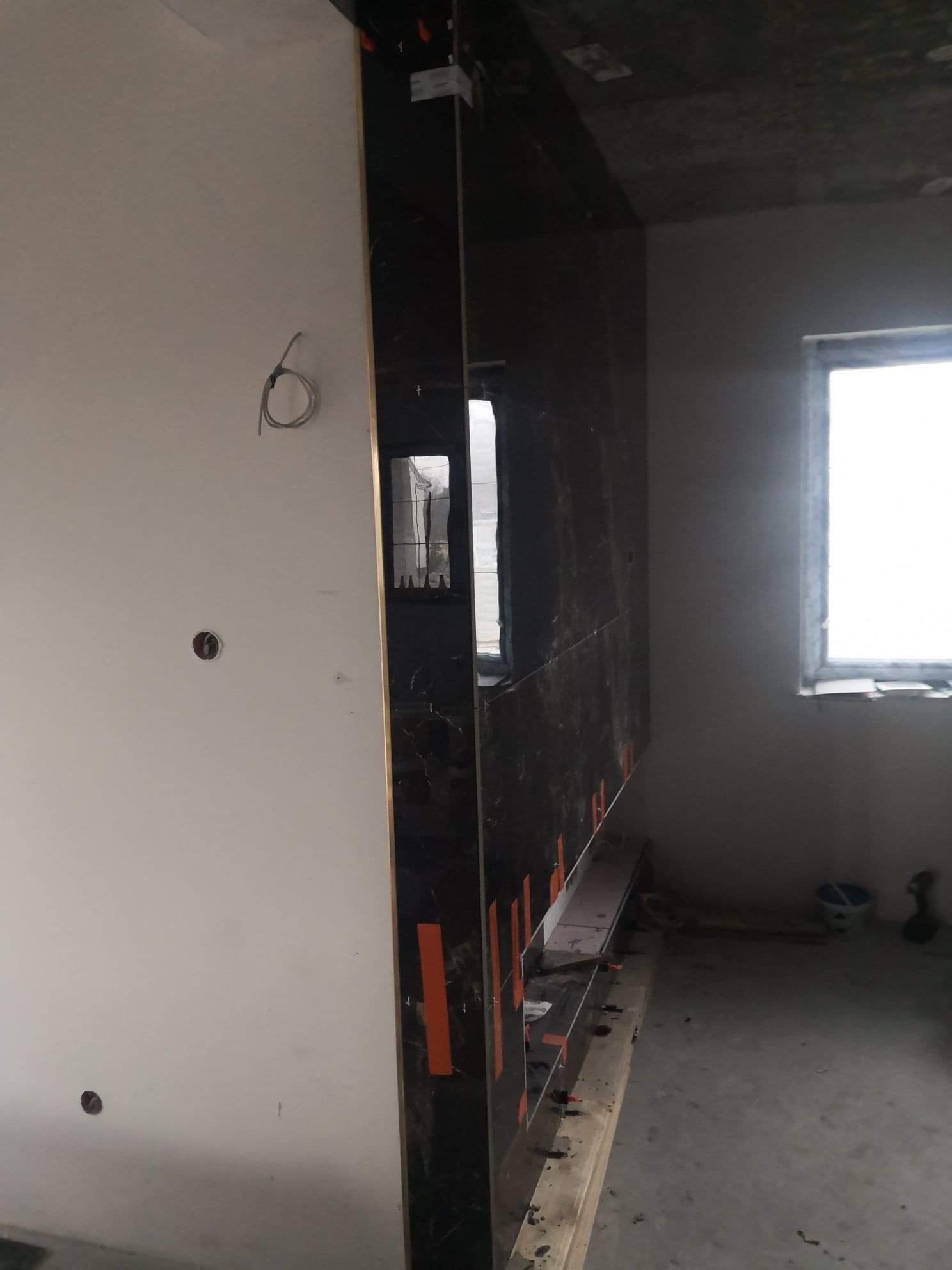 Wnętrze w trakcie prac wykończeniowych, widoczna ściana z czarnych płyt z marmurowym wzorem, częściowo zamontowana, z pomarańczowymi krzyżykami dystansowymi, surowy tynk na ścianach, niedokończony...