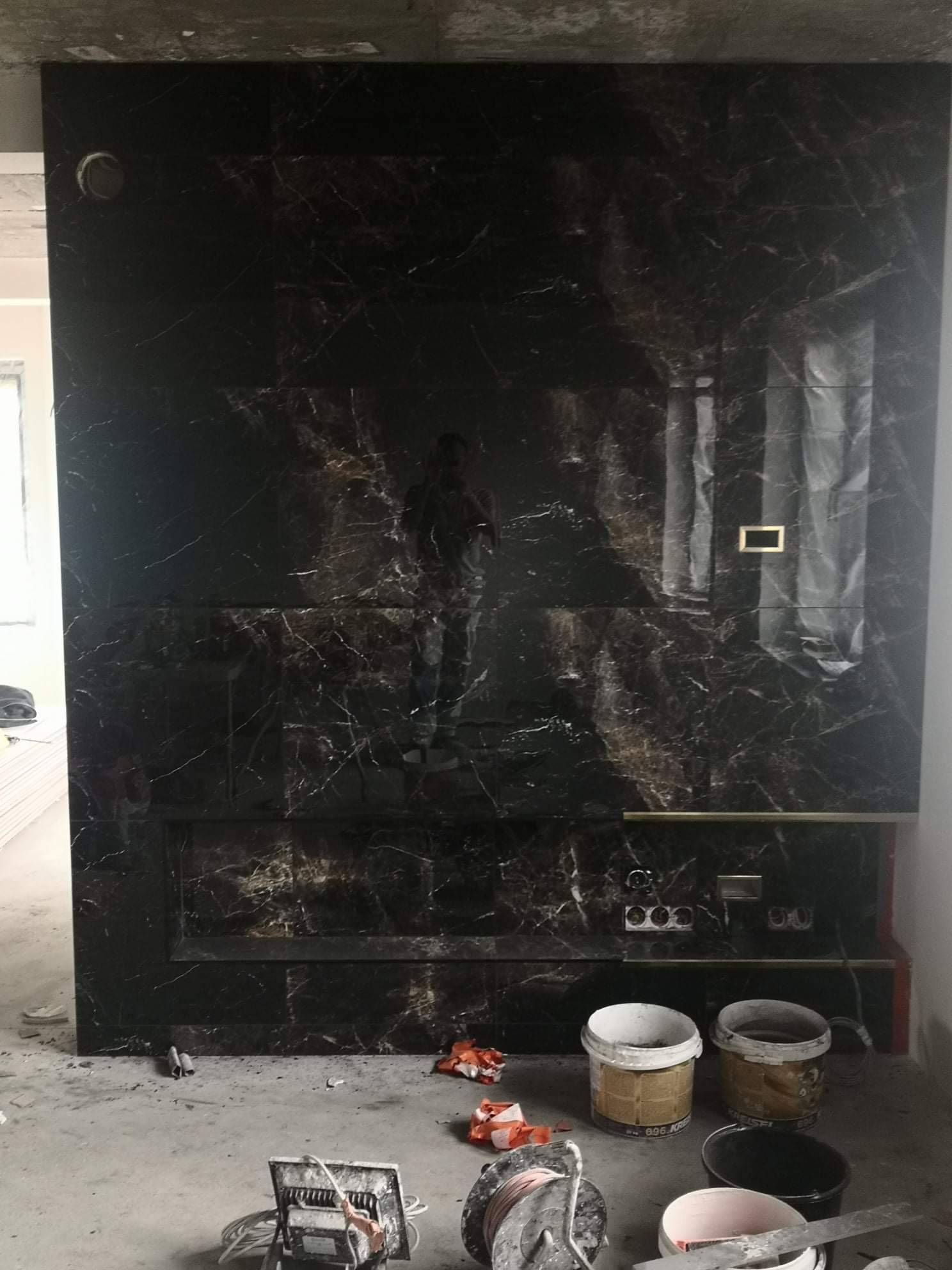 Ciemna ściana z marmurowym wzorem w trakcie wykończenia, widoczne puszki elektryczne, reflektor i puste wiadra po farbie na surowym betonie