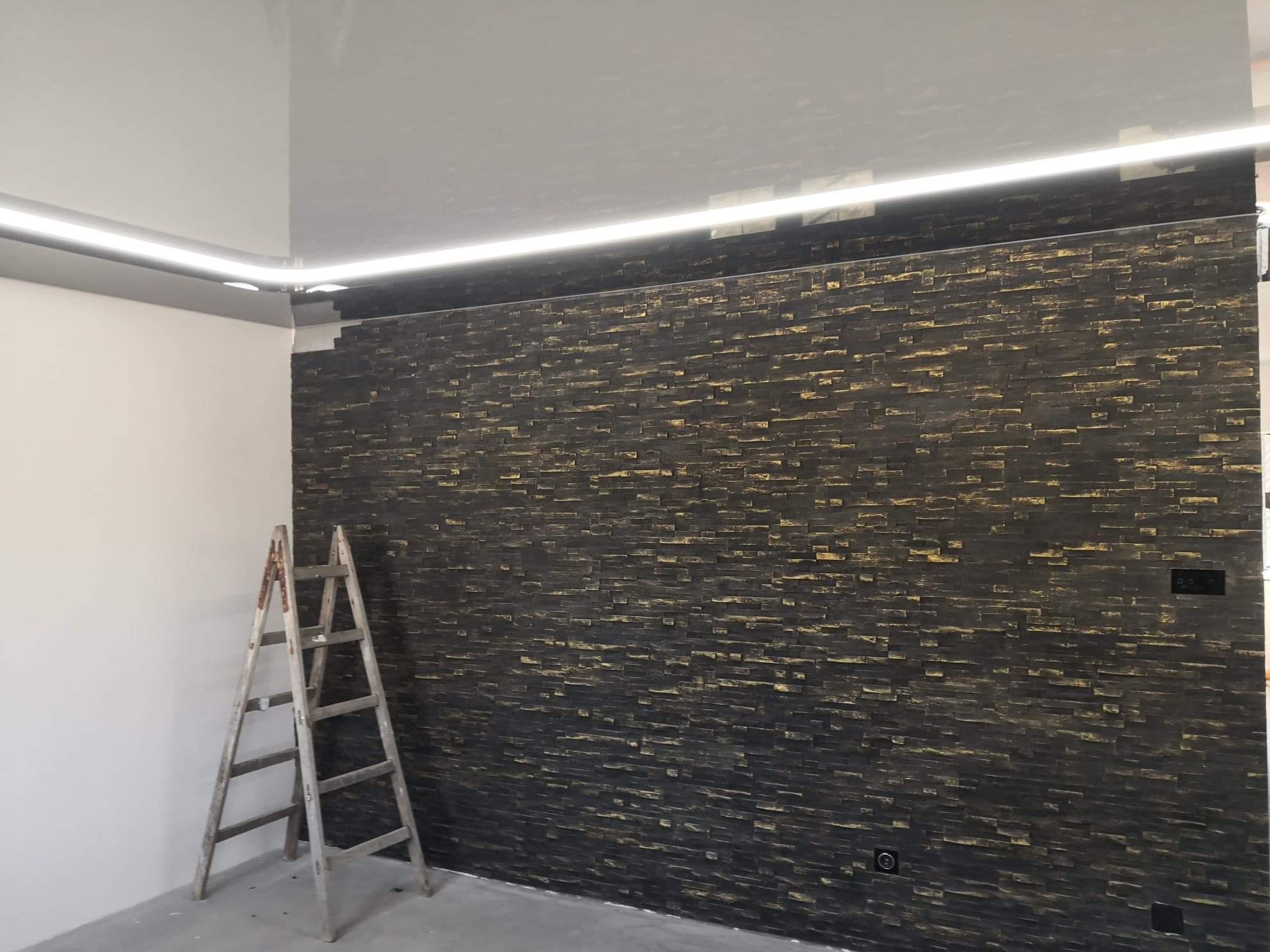 Ściana wykończona dekoracyjną, czarno-złotą okładziną kamienną, widoczna drabina malarska, biały sufit z oświetleniem LED, gniazdka elektryczne na ścianie.