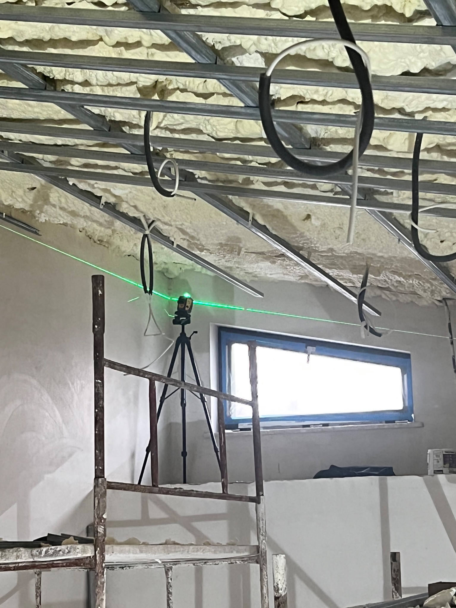 Ocieplony strop z metalową konstrukcją podwieszaną, widoczny laser krzyżowy na statywie i fragment rusztowania, okno w tle.