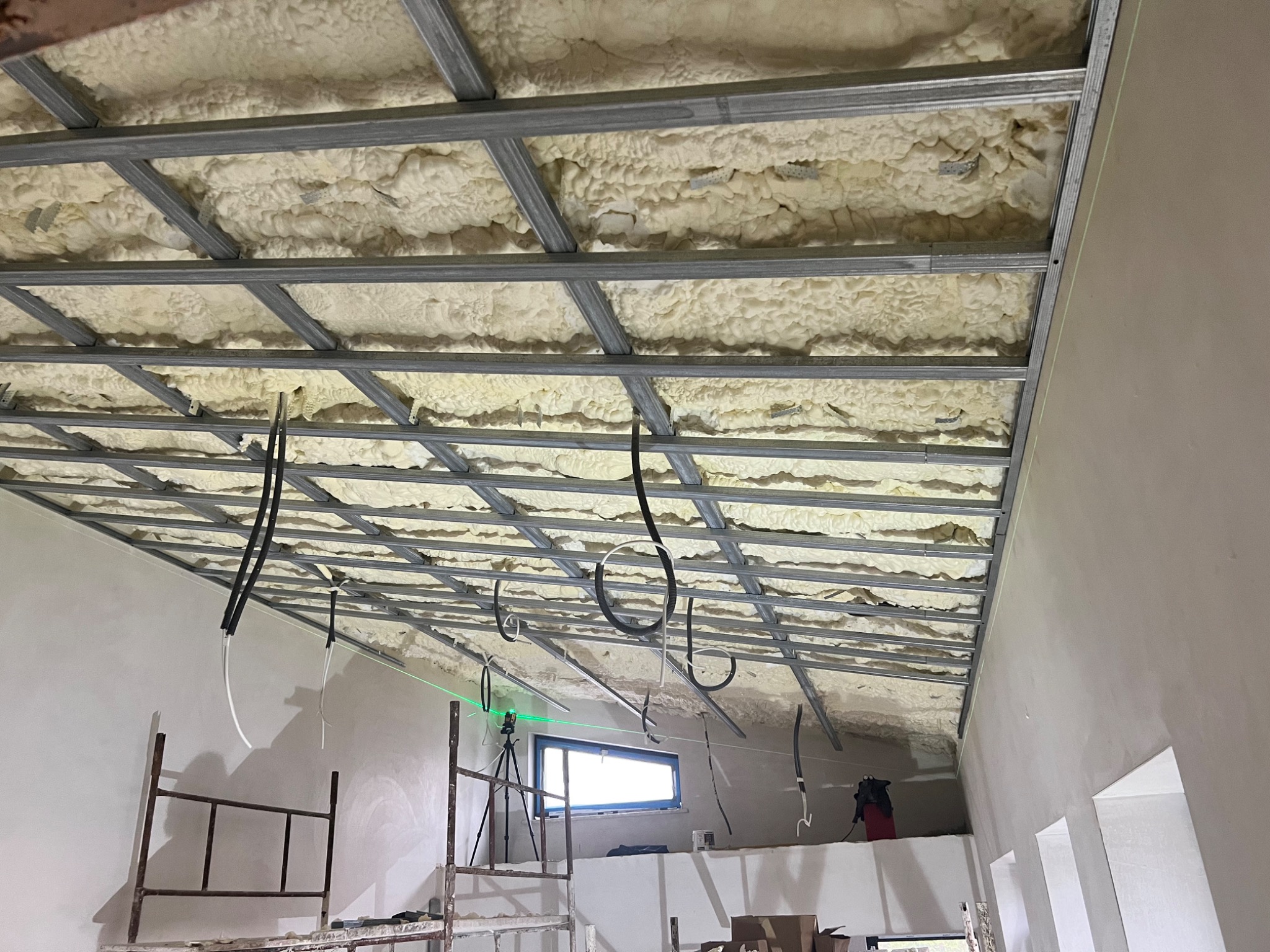 Ocieplenie poddasza pianą PUR widoczne przez metalowy stelaż, na ścianie widoczna poziomica laserowa, w tle okno i rusztowanie.
