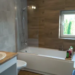 Nowoczesna łazienka z wanną za szklanym parawanem, umywalką na drewnianym blacie, szarymi płytkami na ścianie i oknem z widokiem na zieleń.