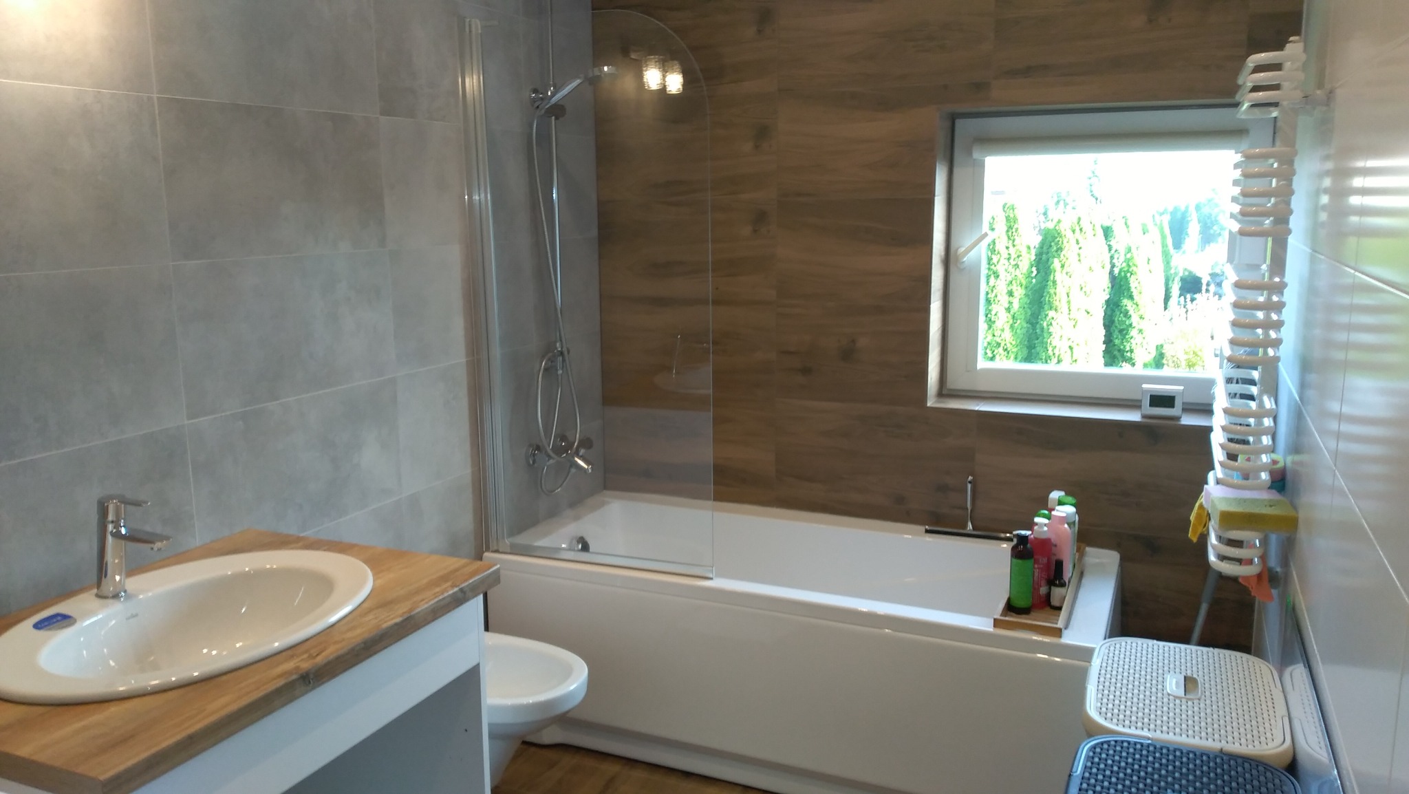 Nowoczesna łazienka z wanną za szklanym parawanem, umywalką na drewnianym blacie, szarymi płytkami na ścianie i oknem z widokiem na zieleń.