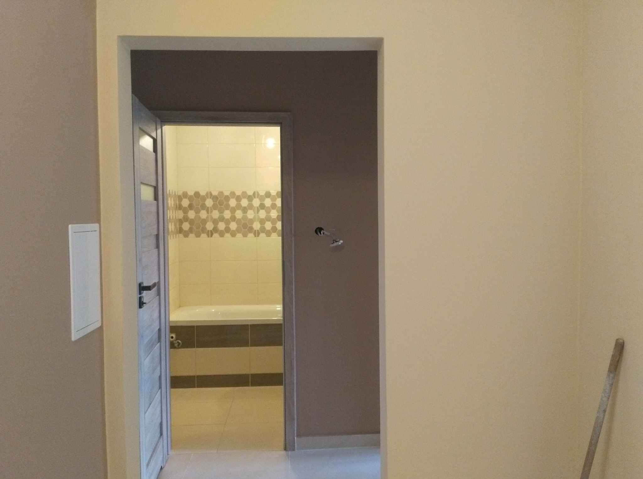 Widok na wykończoną łazienkę przez otwarte drzwi, z widocznymi fragmentami pomalowanych ścian w kolorach beżowym i szarym oraz narzędziem opartym o ścianę.