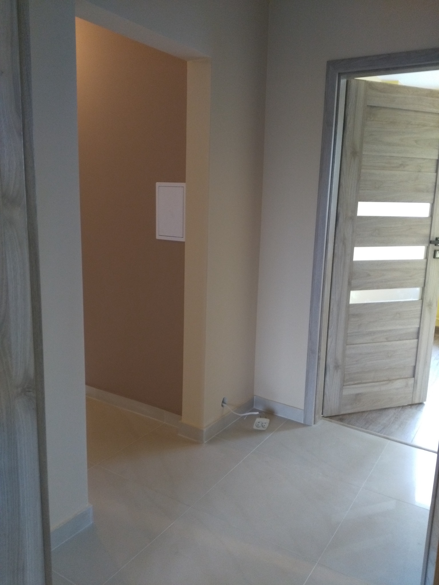 Świeżo pomalowane wnętrze w Łodzi z beżowymi ścianami, jasnymi płytkami podłogowymi i drewnianymi drzwiami z przeszkleniami, widoczna puszka instalacyjna na ścianie.