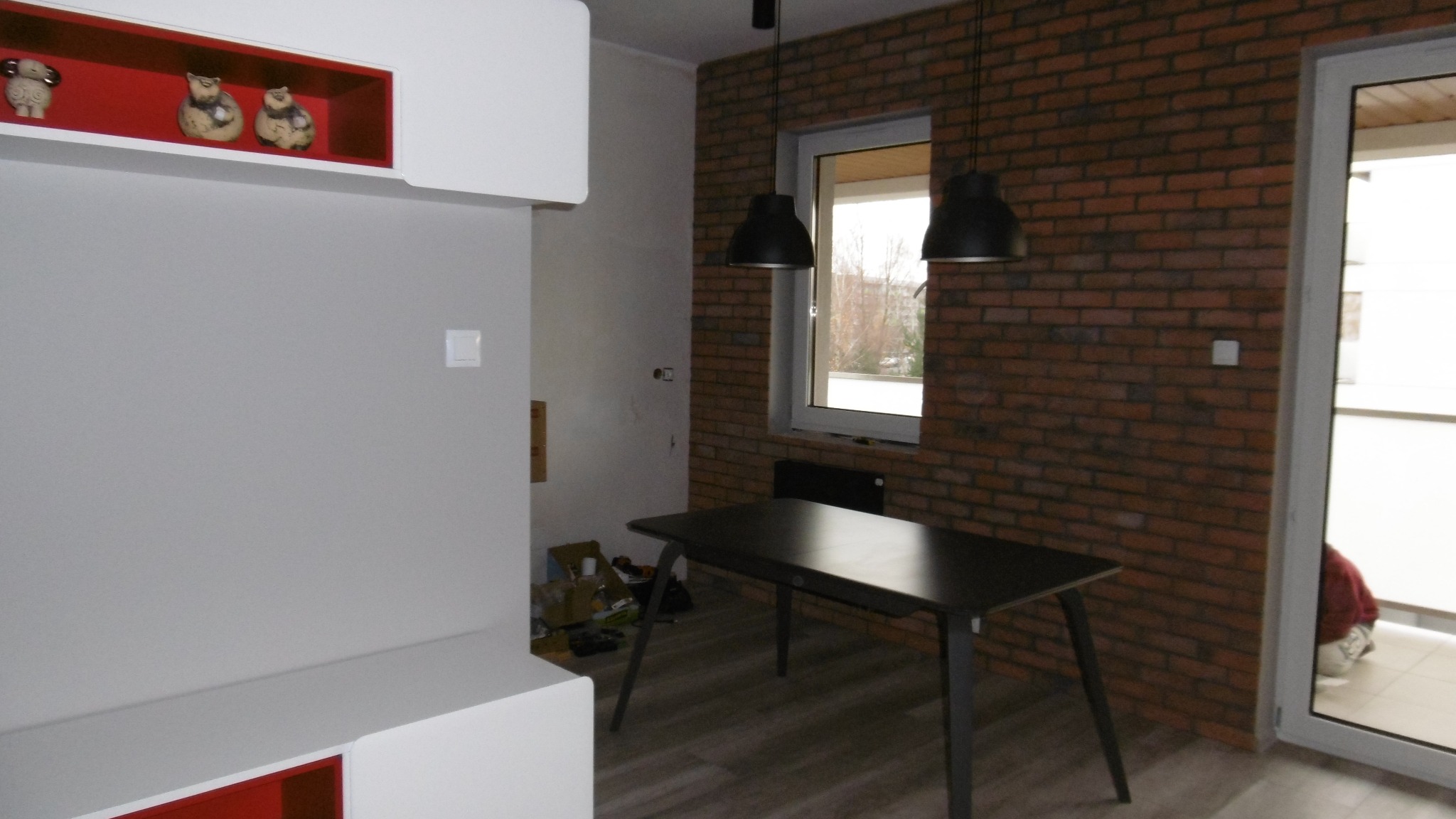 Pokój po remoncie z częściowo odsłoniętą ceglaną ścianą, wiszącymi lampami i nowoczesnymi meblami w kolorze białym i czerwonym.