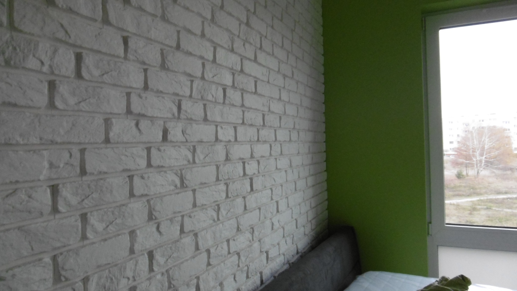 Ściana z białej cegły dekoracyjnej w pokoju, widoczny fragment szarej sofy i zielona ściana obok okna z widokiem na budynki.