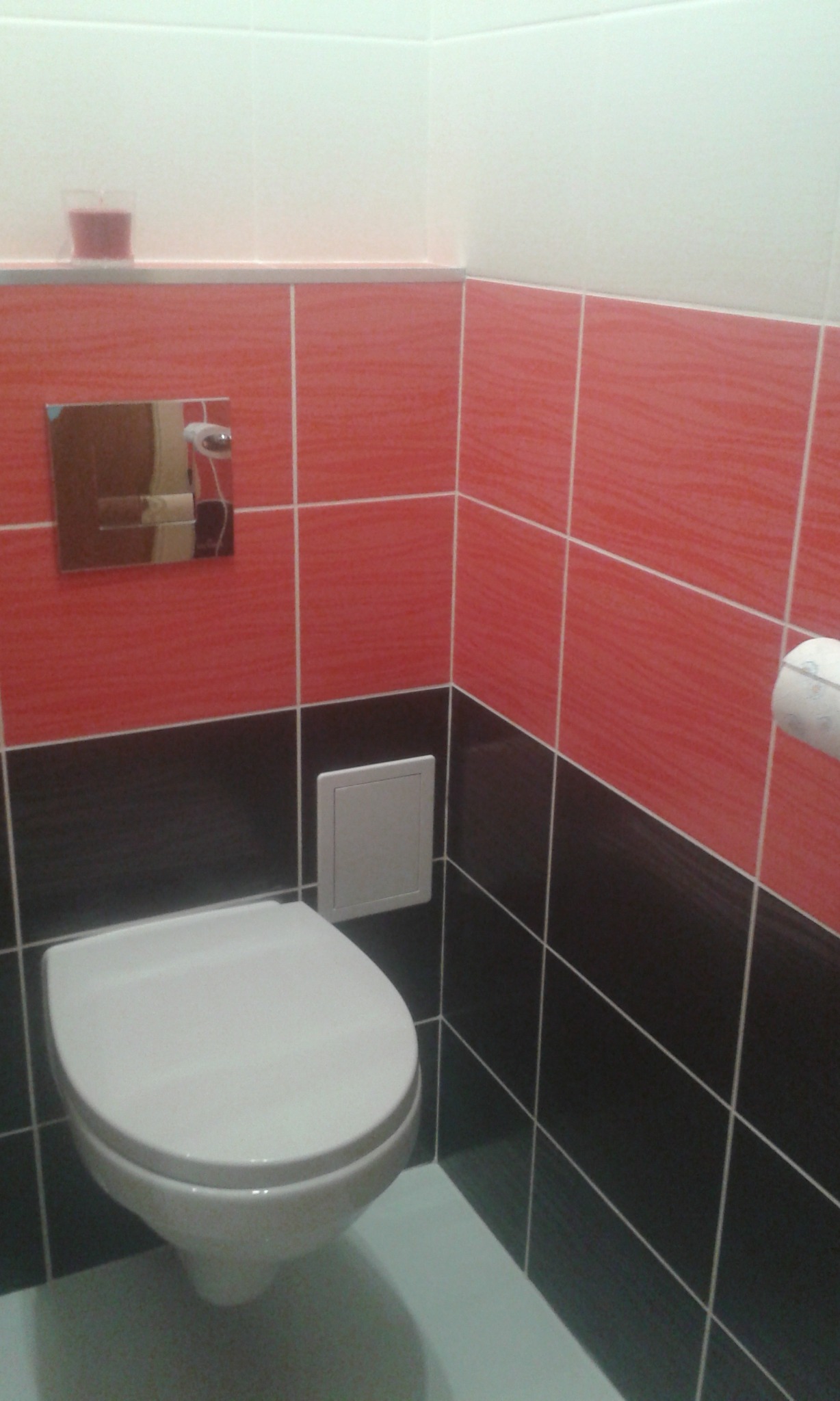 Toaleta wisząca w łazience wykończonej płytkami w kolorach czerwieni i czerni, z widocznym fragmentem ściany z białą półką i świeczką.