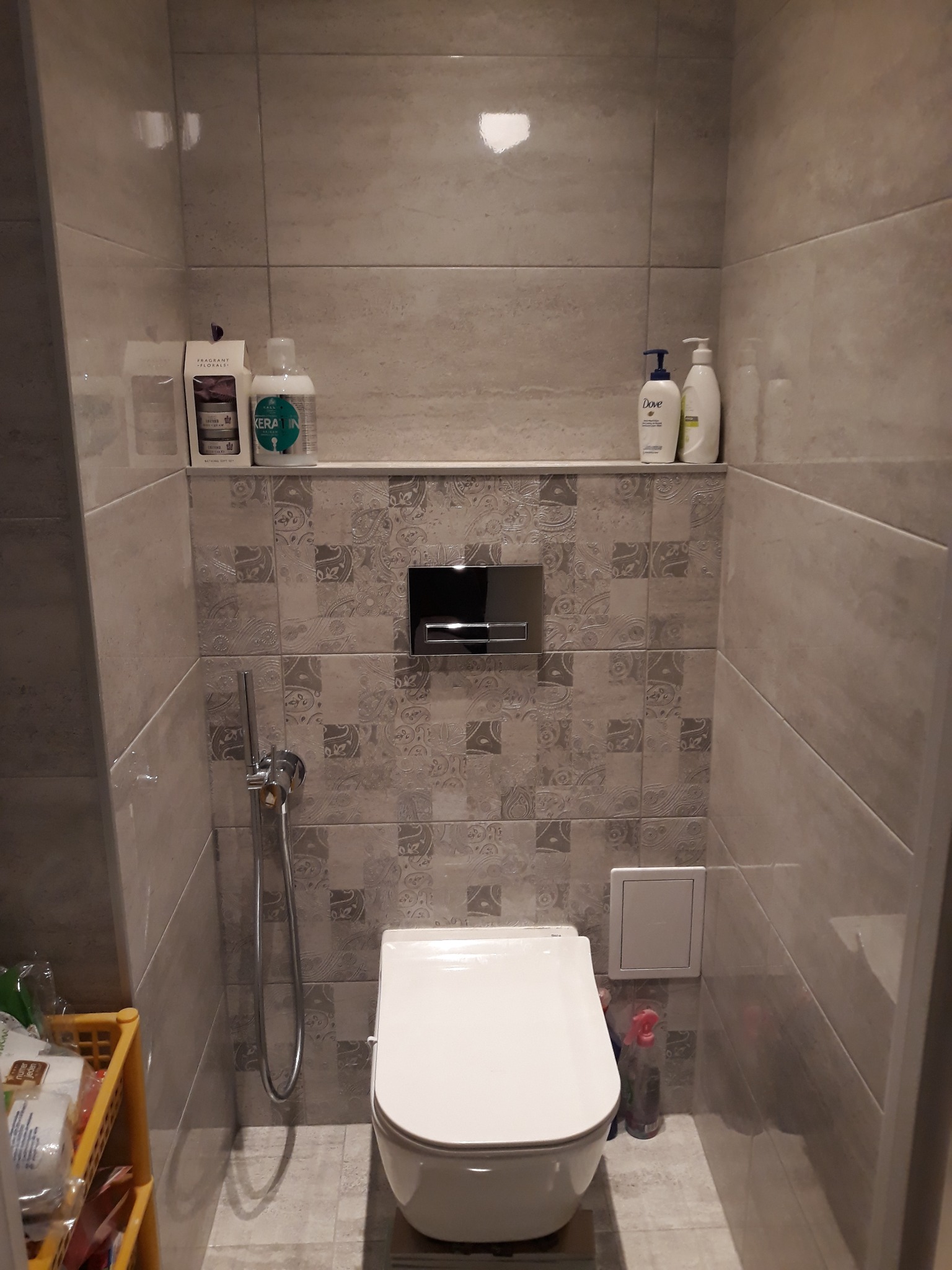 Kompaktowa toaleta w szaro-beżowej łazience z wzorzystymi płytkami, chromowanym panelem sterowania spłuczką i półką z kosmetykami oraz armaturą.