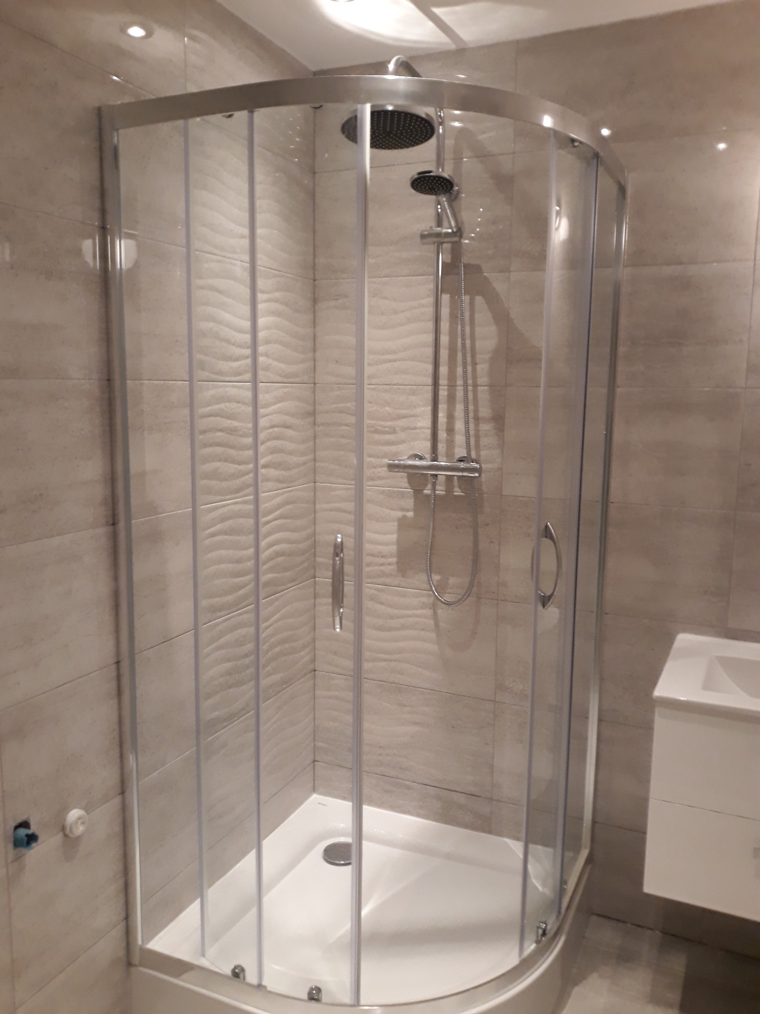 Zamknięta kabina prysznicowa z hydromasażem i deszczownicą, widoczna na tle beżowych płytek o falistej strukturze, z umywalką z szafką po prawej stronie.