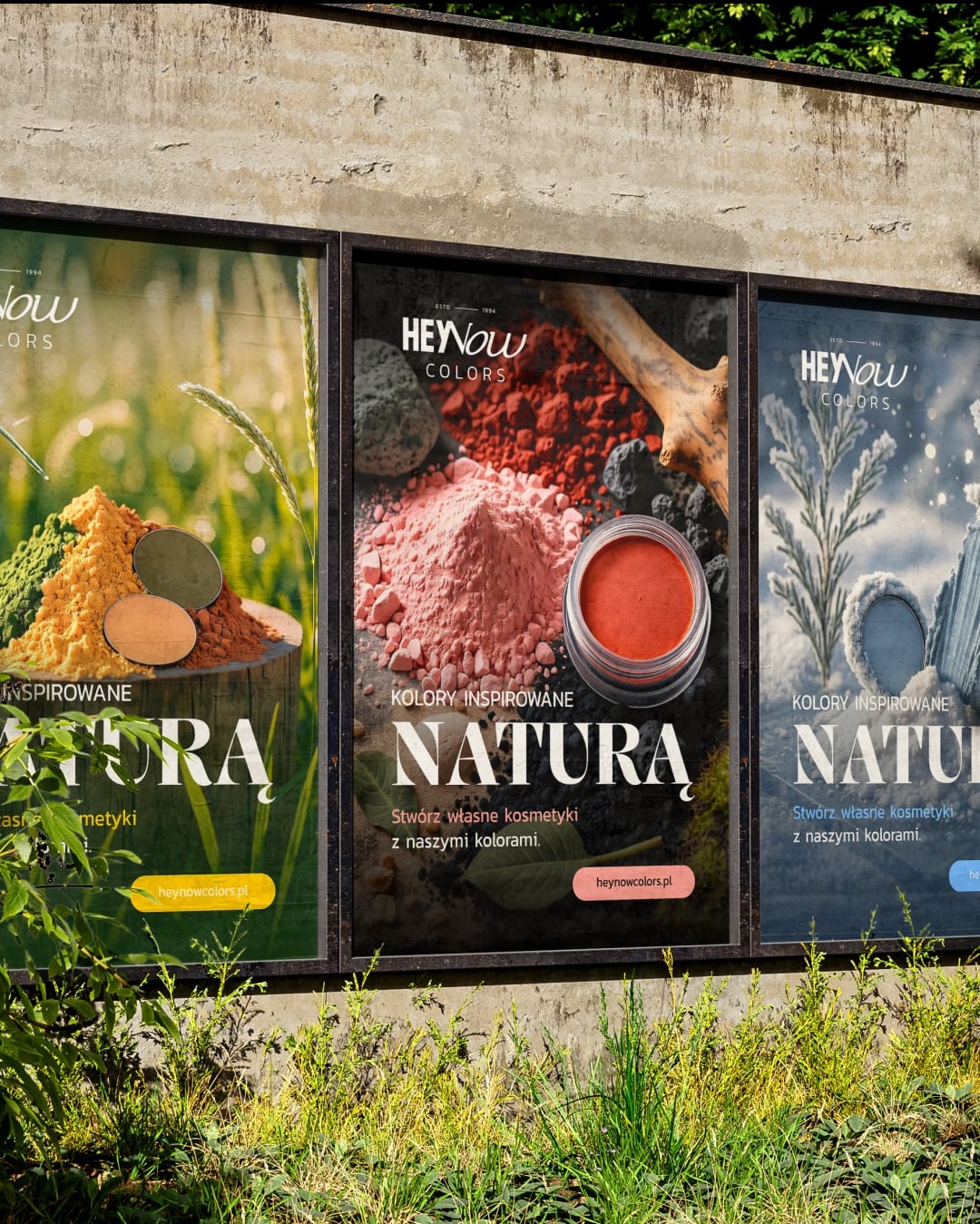 Trzy plakaty reklamowe marki kosmetycznej 'HeyNow Colors' na betonowej ścianie, z motywami natury i kolorowymi proszkami. Reklama zachęca do tworzenia własnych kosmetyków.