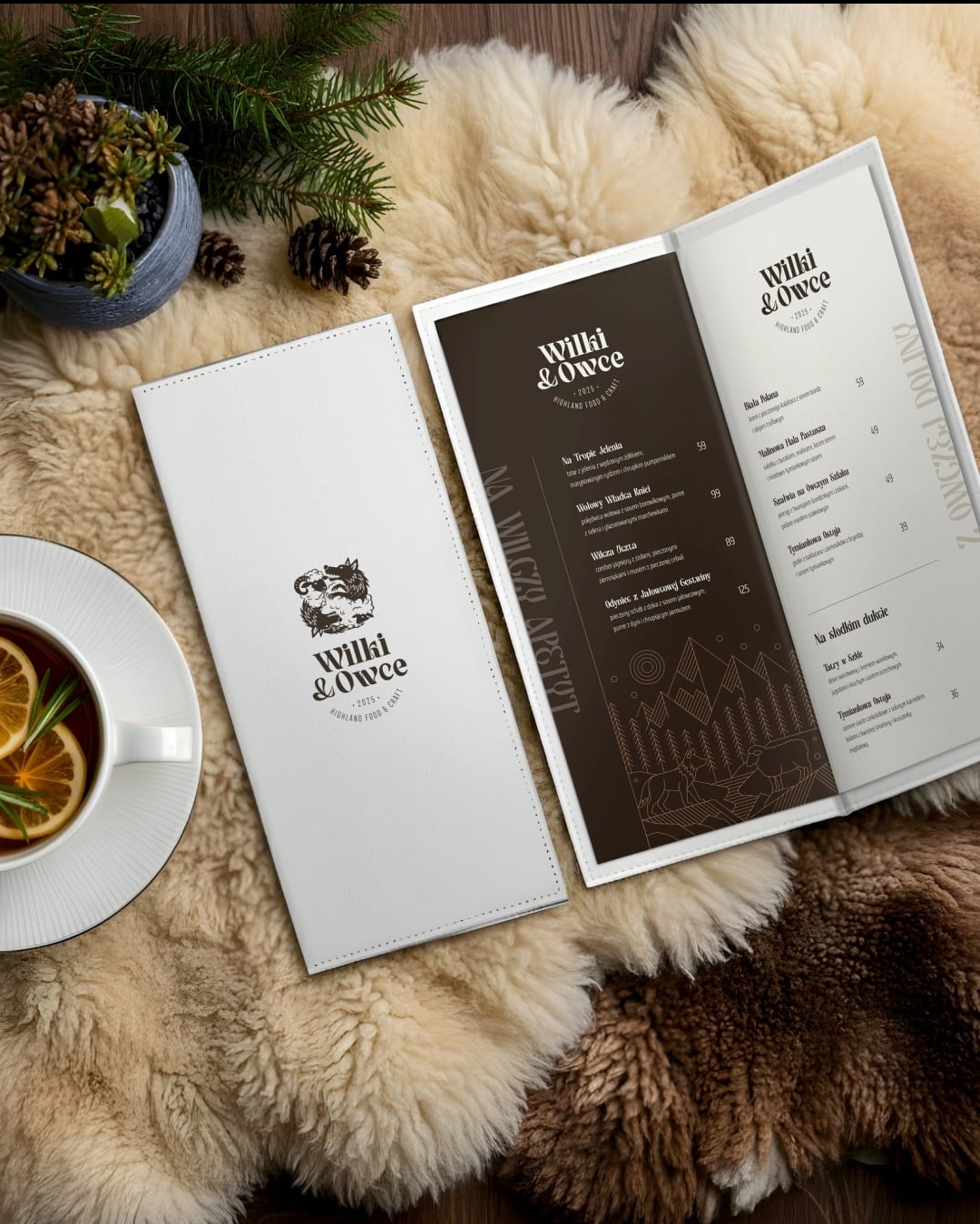 Menu restauracji Wilki & Owce z logo na okładce, otwarte na stronie z daniami, obok filiżanka herbaty i dekoracje świąteczne na futrzanym tle.