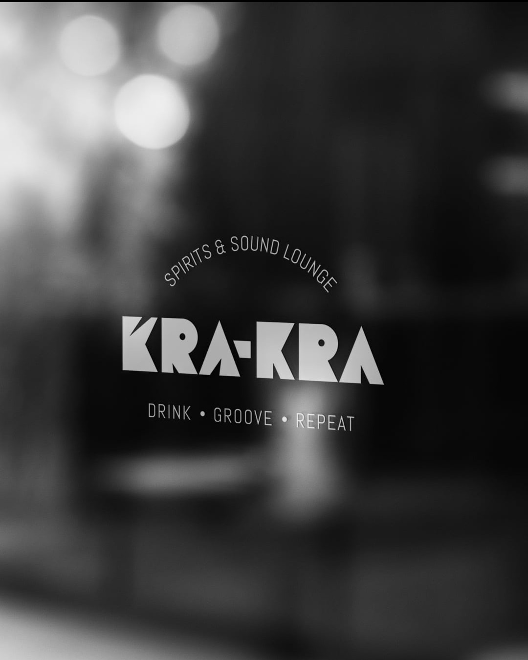 Logo 'KRA-KRA Spirits & Sound Lounge' w bieli na ciemnym tle, widoczne przez szybę z rozmytymi światłami w tle. Napis: Drink, Groove, Repeat.