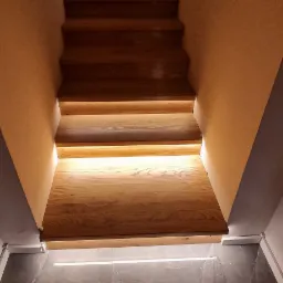 Drewniane schody z podświetleniem LED, widok z dołu, szare płytki podłogowe, suszarka na ubrania i obraz w tle.