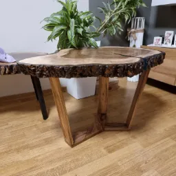 Stolik kawowy z blatem z naturalnego drewna o nieregularnym kształcie krawędzi, z żywicą epoksydową w szczelinach, na drewnianych nogach w kształcie litery V, obok rośliny doniczkowej i szmatki...