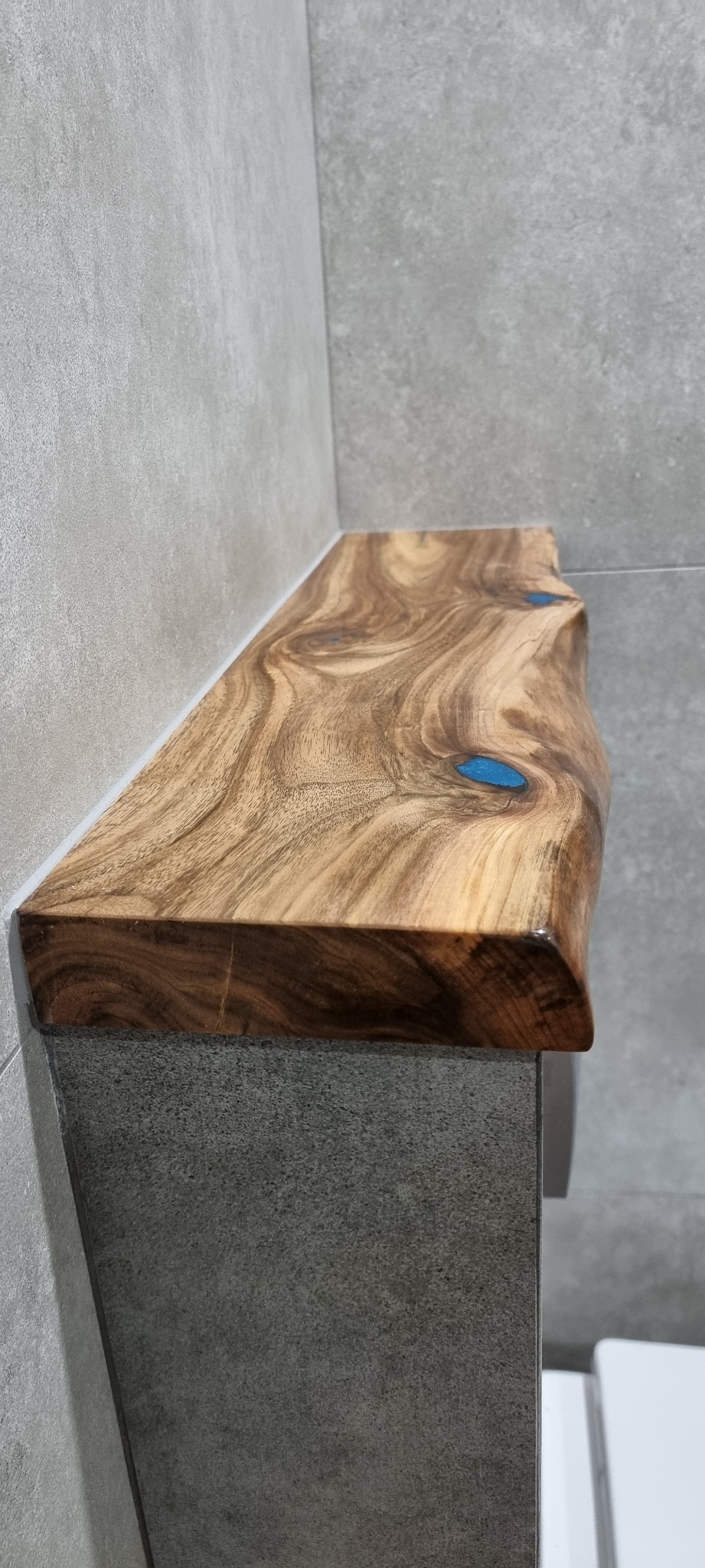 Półka z drewna egzotycznego z żywicą epoksydową w kolorze turkusowym, zamontowana w szarej łazience z betonową fakturą ścian.