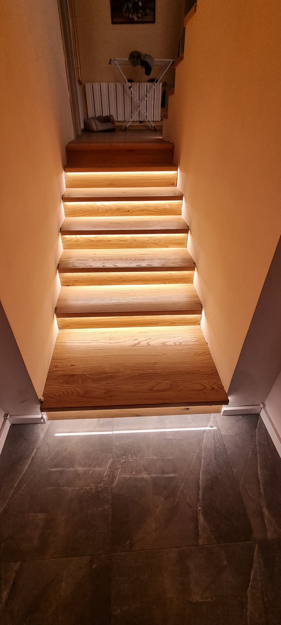 Drewniane schody z podświetleniem LED na każdym stopniu, widok od dołu, szare płytki na podłodze, suszarka na pranie w tle.