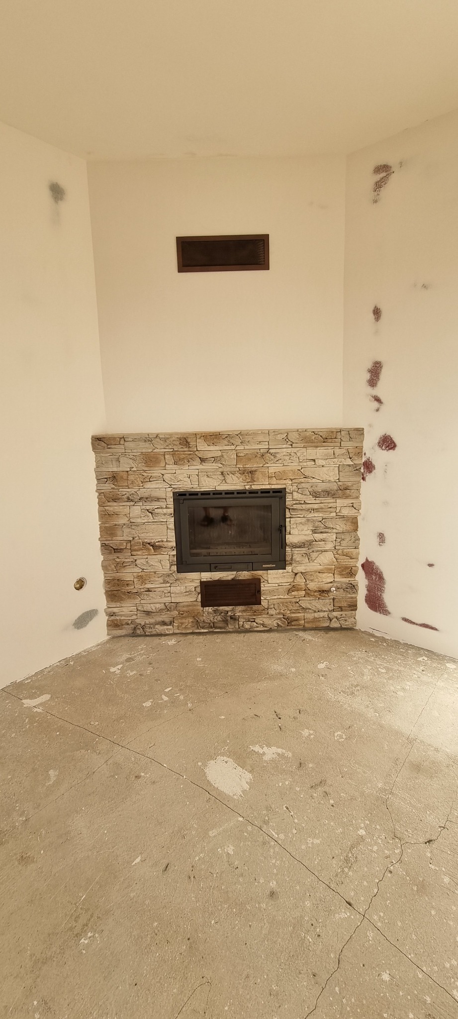 Obudowa kominka wyłożona kamieniem dekoracyjnym, surowy beton na podłodze, ślady po remoncie na ścianach, kratka wentylacyjna nad kominkiem.