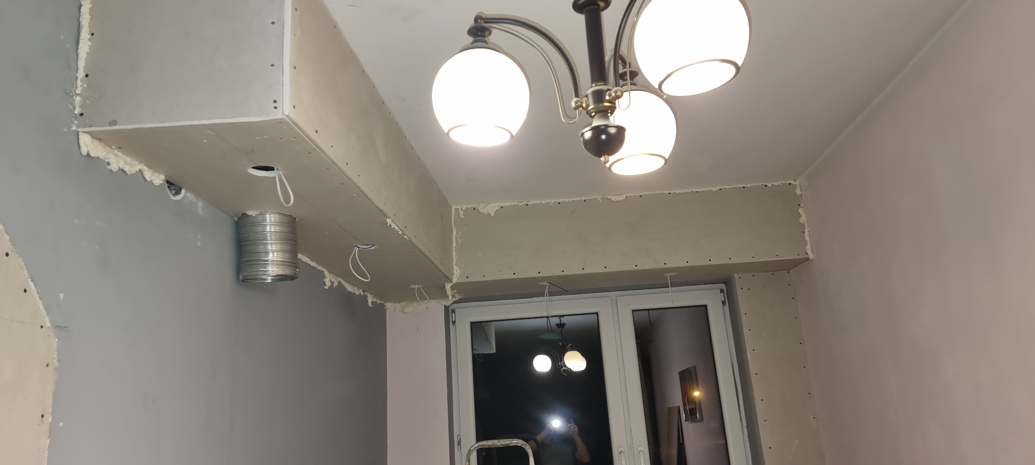 Sufit podwieszany z płyt karton gips, widoczne profile, wata szklana w szczelinach, kabel wystający z otworu, metalowy przewód wentylacyjny, lampa sufitowa w tle okna.