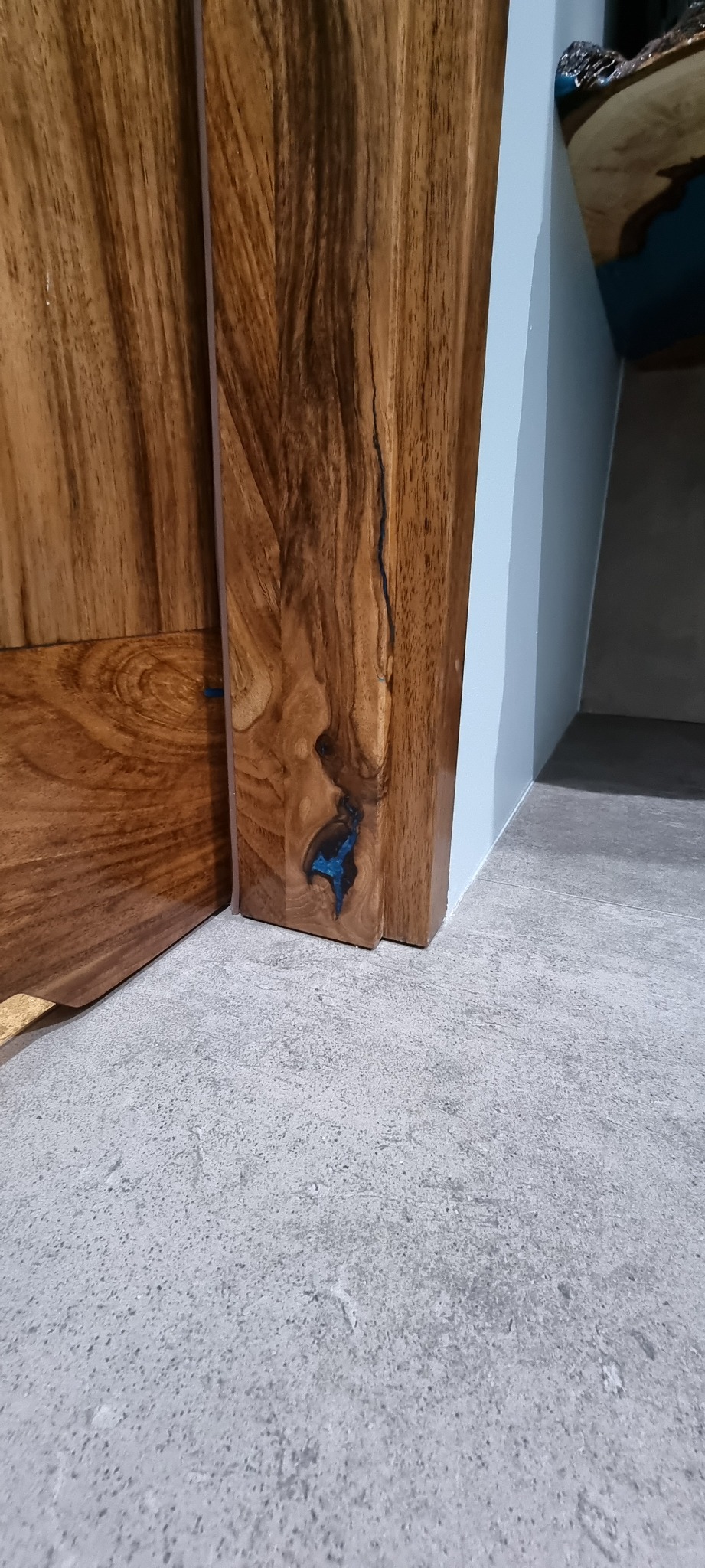 Fragment blatu drewnianego z żywicą epoksydową w kolorze turkusowym, przylegający do szarej ściany i betonowej podłogi, widoczny z niskiej perspektywy.