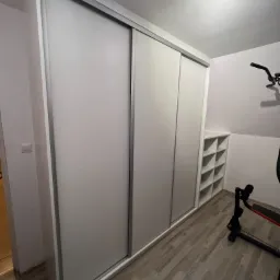 Biała szafa przesuwna na wymiar z aluminiowymi ramami, w pokoju na poddaszu ze skośnym sufitem, obok regał z kwadratowymi półkami i sprzęt do ćwiczeń.