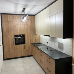 Nowoczesna kuchnia z drewnianymi frontami szafek, czarnym blatem i piekarnikiem w zabudowie, oświetlona wiszącą lampą w kształcie sześcianu.