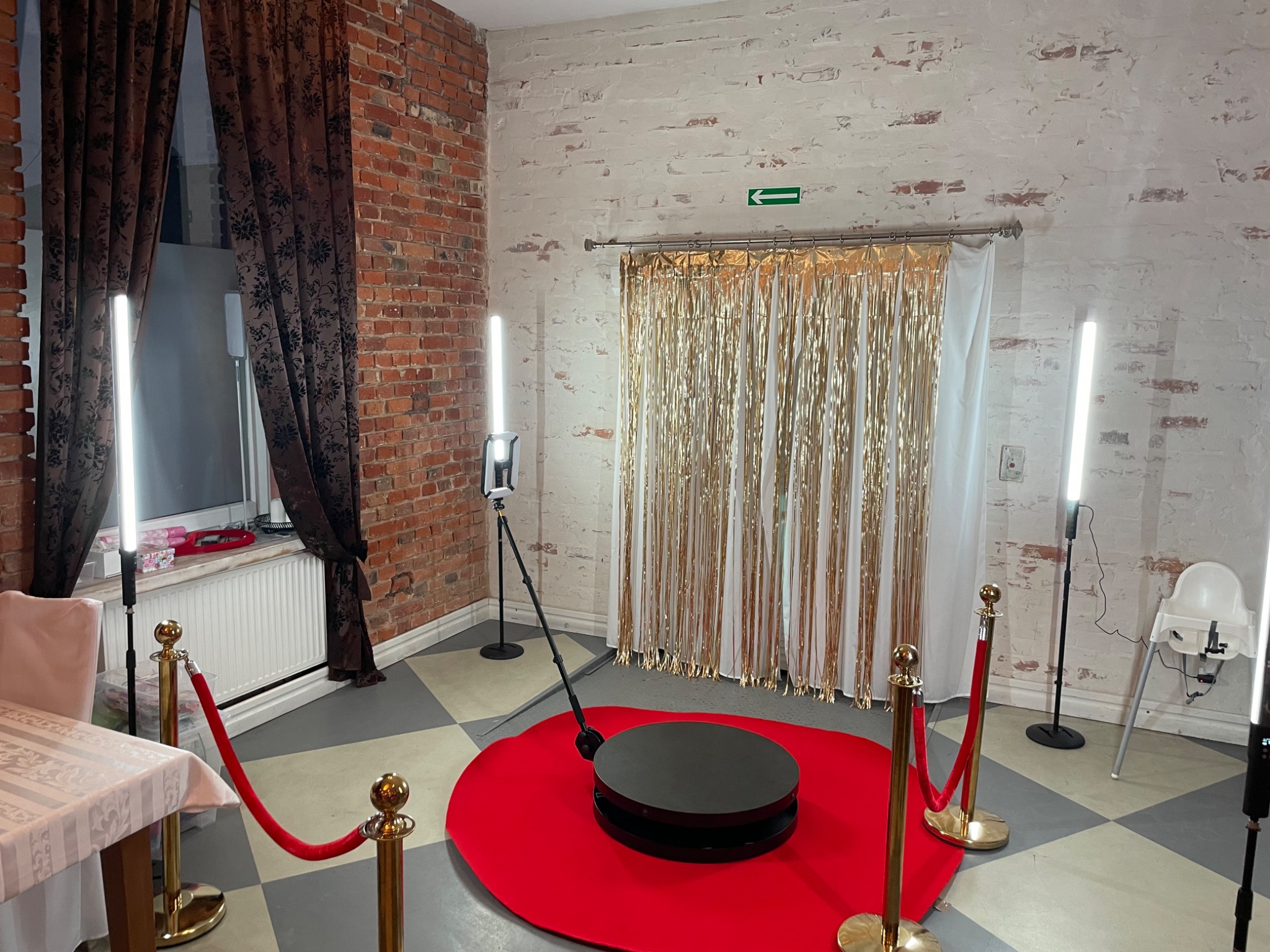 Przygotowana przestrzeń do zdjęć z obrotową platformą na czerwonym dywanie, otoczona złotymi słupkami z czerwonymi sznurami, lampami LED i złotą kurtyną na tle ceglanej i malowanej ściany.