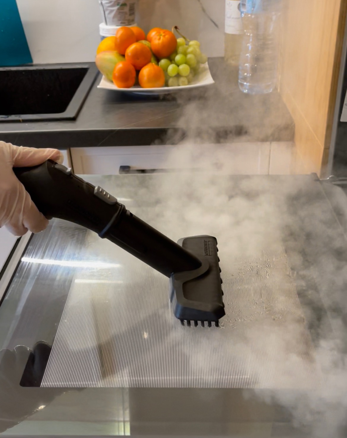 Czyszczenie parowe płyty grzewczej w kuchni, widoczna ręka w rękawicy ochronnej i dysza parownicy, w tle owoce na blacie.