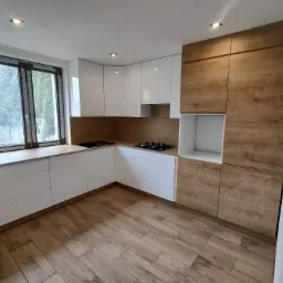 Nowoczesna kuchnia z białymi szafkami i drewnianymi akcentami, widok na okno z widokiem na zewnątrz, drewnopodobna podłoga, oświetlenie punktowe.