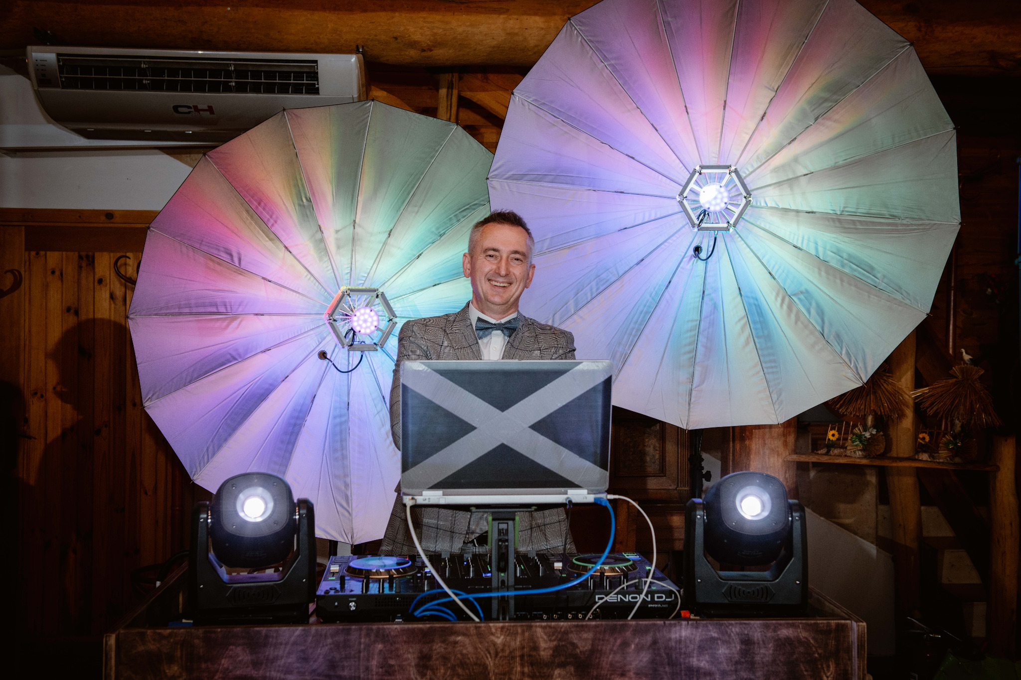 Uśmiechnięty DJ w muszce i marynarce w kratę, za konsolą Denon DJ, otoczony kolorowymi parasolami-lampami, z laptopem z naklejką w kształcie litery X na tle drewnianej ściany.
