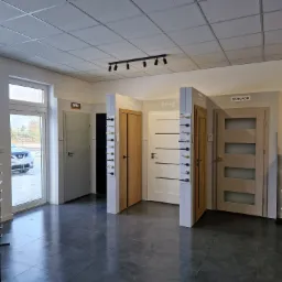 Ekspozycja modeli drzwi różnych marek (DRE, Porta, Erkado) w salonie, z prezentacją klamek na stojakach i wbudowanych w ścianki działowe, widoczne okno wystawowe z samochodem na zewnątrz.