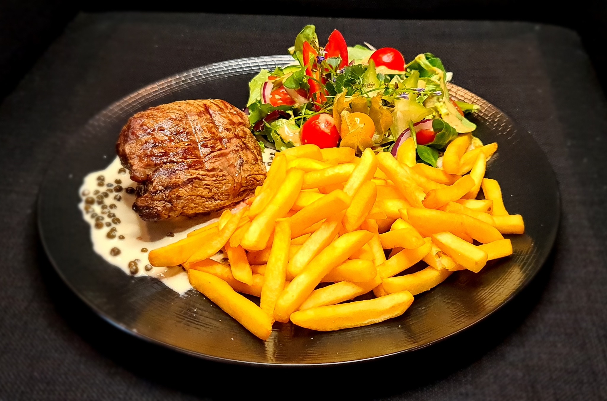 Stek wołowy z sosem pieprzowym, frytkami i sałatką z pomidorkami cherry oraz physalis na czarnym talerzu.