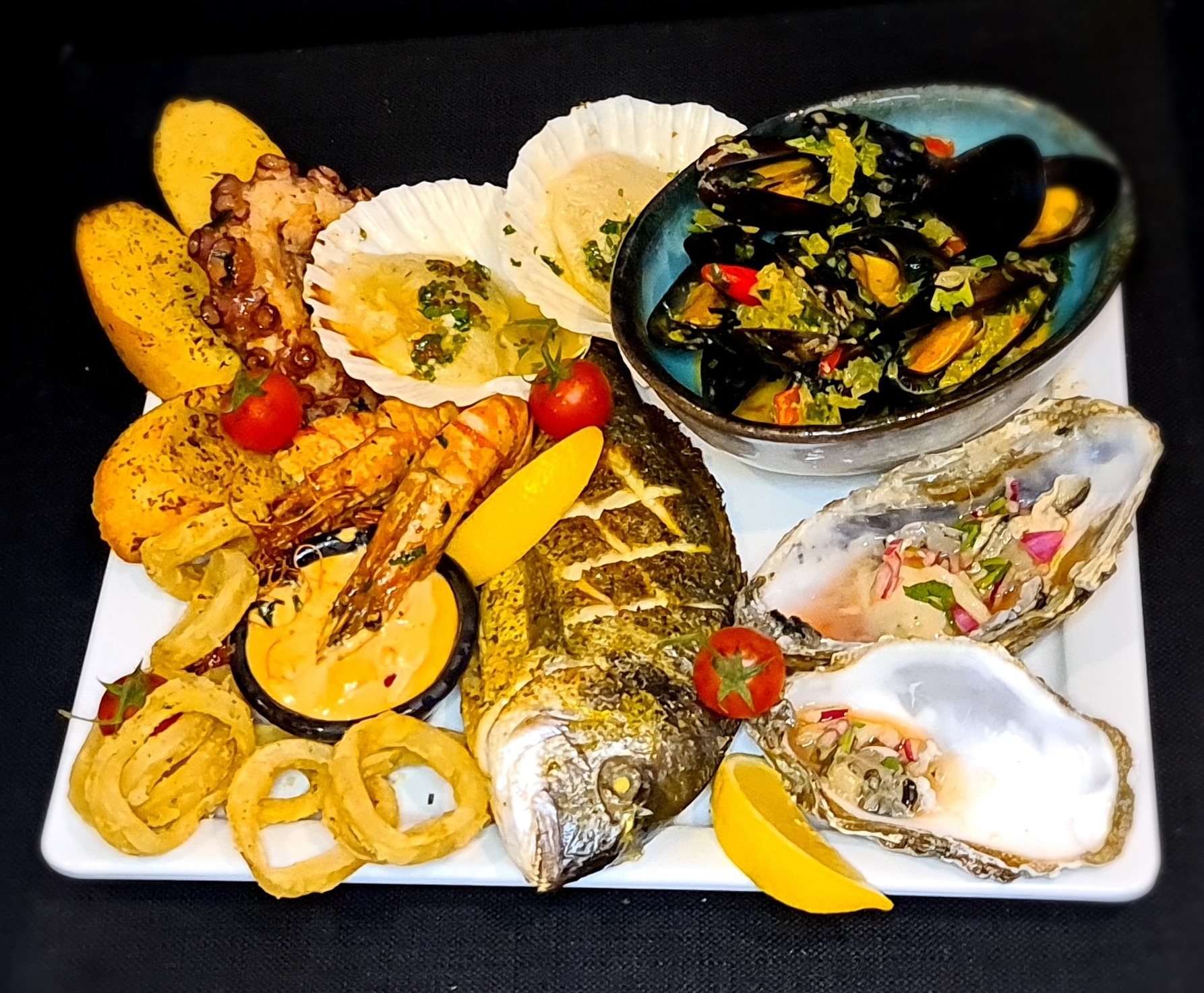 Elegancko podane owoce morza na białym talerzu: grillowana ryba, ostrygi, małże w sosie, kalmary w cieście, krewetki, przegrzebki, ziemniaki i pomidorki koktajlowe.