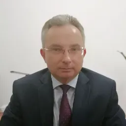 Portret biznesowy mężczyzny w okularach i garniturze, na neutralnym tle, z delikatnym uśmiechem.