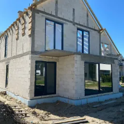 Surowy stan domu jednorodzinnego w Warszawie z zamontowanymi oknami w czarnych ramach i widoczną konstrukcją dachu w trakcie budowy.