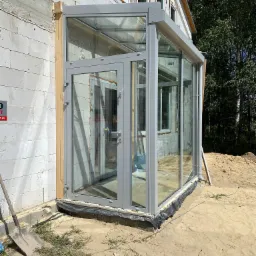 Szklana weranda z szarymi ramami, dobudowana do surowego budynku z białego bloczka, widoczna szpadel i piasek wokół.