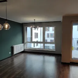 Puste, nowocześnie urządzone wnętrze mieszkania z ciemnozieloną ścianą, drewnianą podłogą, grzejnikiem, oknem z widokiem na budynek i designerskimi lampami wiszącymi.